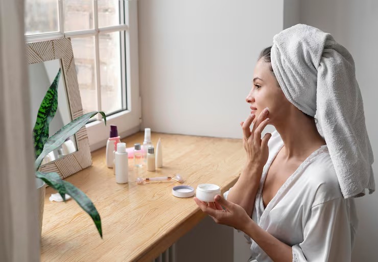Woman in white robe applying face cream by window, surrounded by skincare products